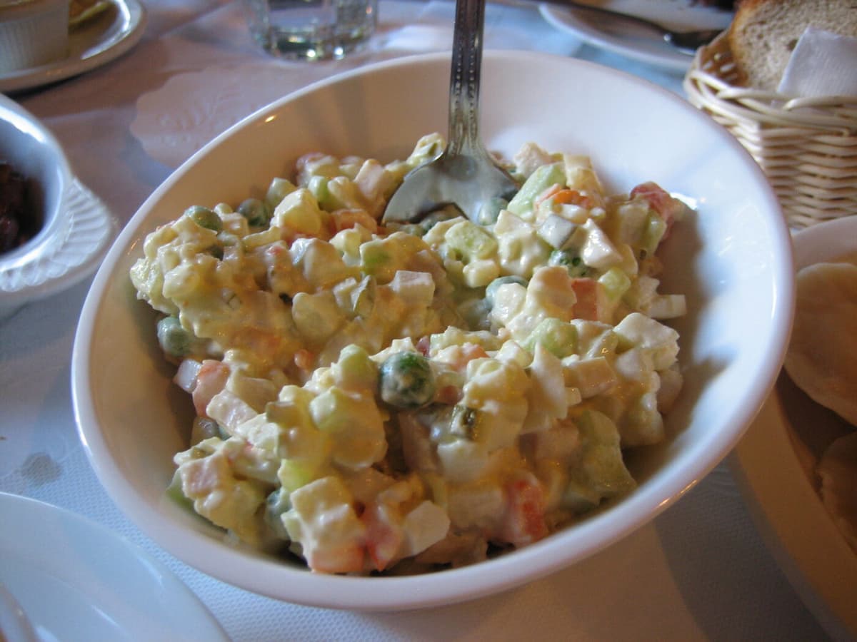A white bowl of Olivier salad (Russian potato salad) with diced potatoes, peas, carrots, egg, and pickles in mayonnaise on a set table